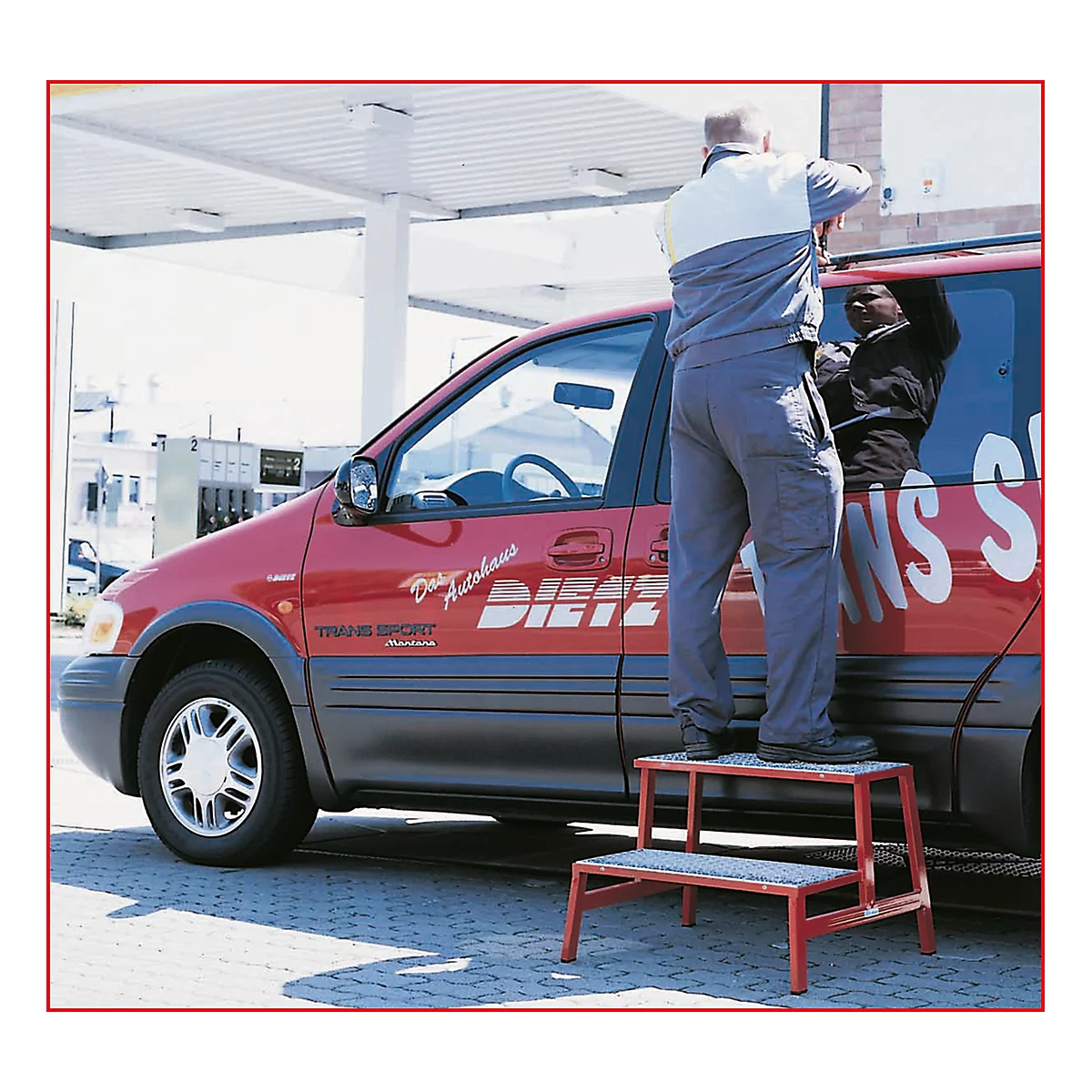 Mann auf roter Trittleiter, repariert rotes Auto. Auf dem Auto steht: "Diez Autohaus Trans Sport".